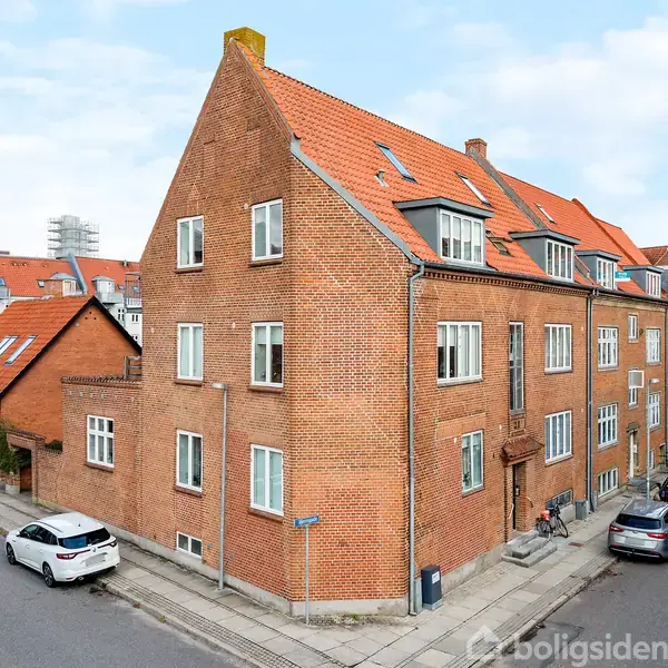 Et rødt murstenshus med tegltag står på et hjørne. Flere vindueskviste og parkerede biler omkranser bygningen i et bymiljø. Skyfri himmel.