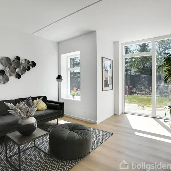 Stue med en mørkegrå sofa, et rundt sofabord med en vase og et stort vindue med udsigt til en have. Lyse vægge og trægulve skaber en moderne atmosfære.