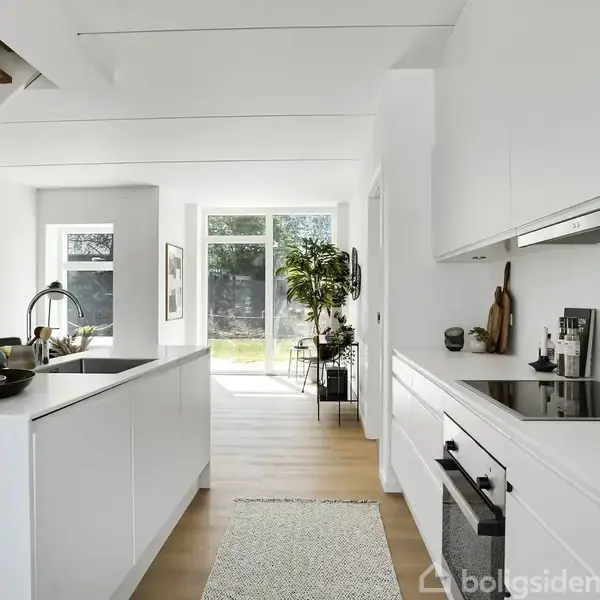 Et moderne køkken med hvide skabe og integrerede apparater. En køkkenø med dekorationer står centralt. I baggrunden er der store vinduer, der giver udsigt til en grøn baghave.
