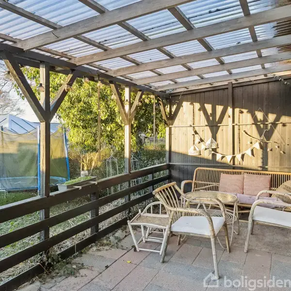 Overdækket terrasse med sofa og stole i flet samt et bord. I baghaven ses en trampolin og frodige grønne buske. Himmel er blå med få skyer.