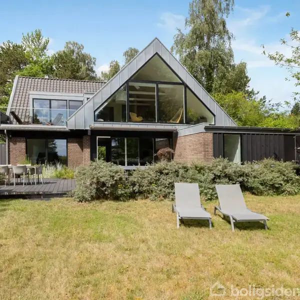 Hus med moderne arkitektur, store glasvinduer og gråt tag, stående i en velplejet have med græsplæne. To grå liggestole er placeret foran huset, omgivet af træer og buske.
