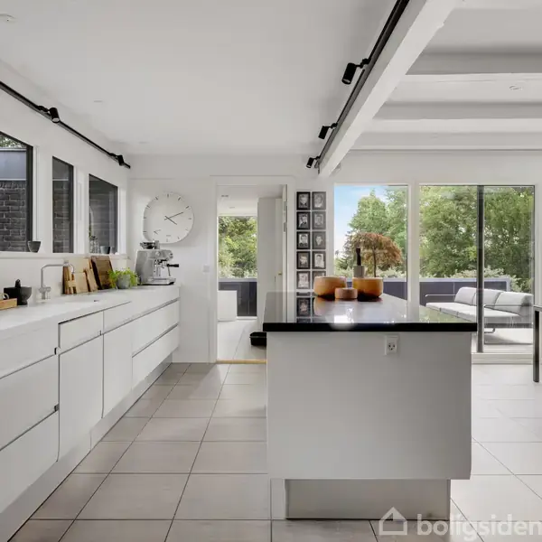 Et moderne køkken med hvide skabe og en central ø i rustfrit stål er beliggende i et lyst rum med store vinduespartier mod en terrasse og have.