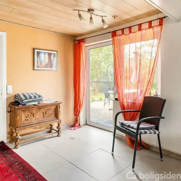 Stol står foran vinduesparti med orange gardiner i et rum med hvidt loft og trægulv. Et lille bord med foldedækkede tekstiler ses. Udsigt til en terrasse udenfor.