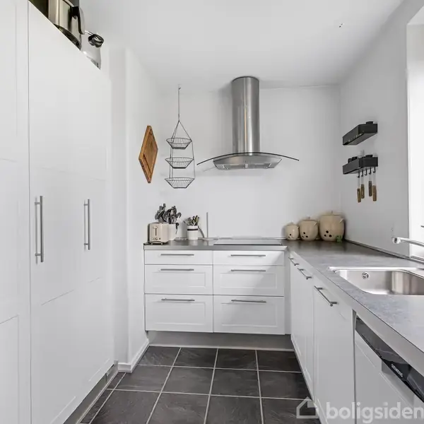 Et moderne køkken med hvide skabe og gråt flisegulv, hvor der er en vask og stål emhætte. Knive og beholdere er placeret på bordet. En stor vindue viser grønne træer udenfor.