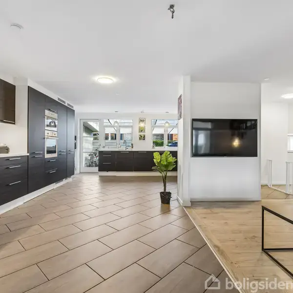 Køkken med mørke skabe strækker sig langs to vægge, mens en stue med en tv-skærm er til højre. Lysindfald fra vinduer og et sofabord med blomster ses.