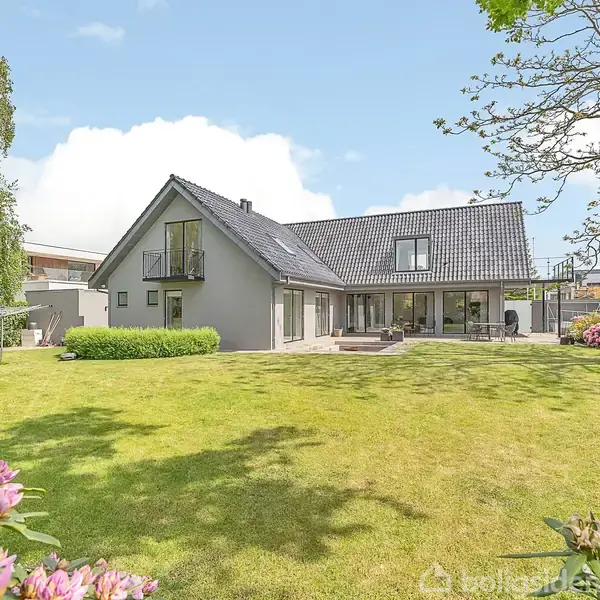 Hus står i en grøn have, omgivet af buske og træer. Det har skråt tag, store vinduer, og en veranda. Atmosfæren er lys og fredfyldt med en blå himmel.