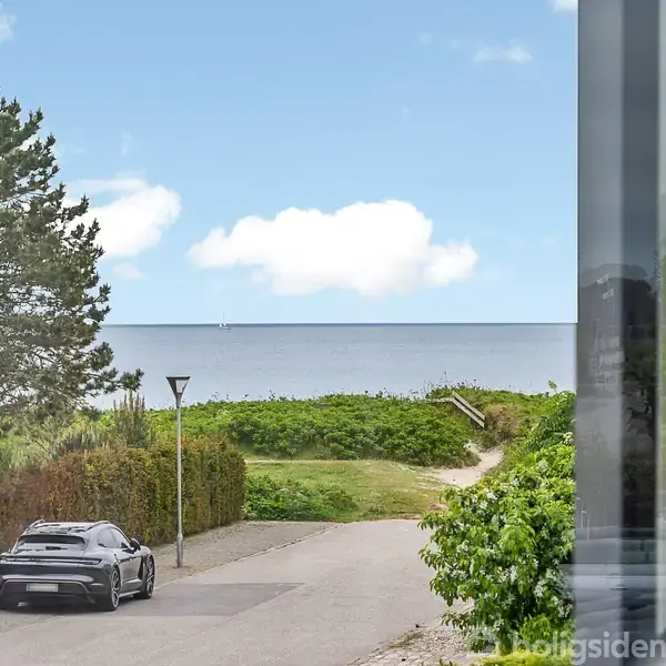 En sort bil holder parkeret på en vej ved siden af et træ. Vejen fører til havet, hvor en lille sti går gennem grønne buske til stranden under en blå himmel med skyer.