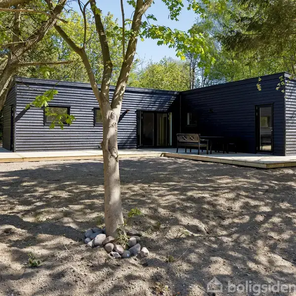 Et sort, moderne hus står stille blandt træer i en skovklædt have. Gruset jord omgiver huset, og terrassen har udendørs møbler.