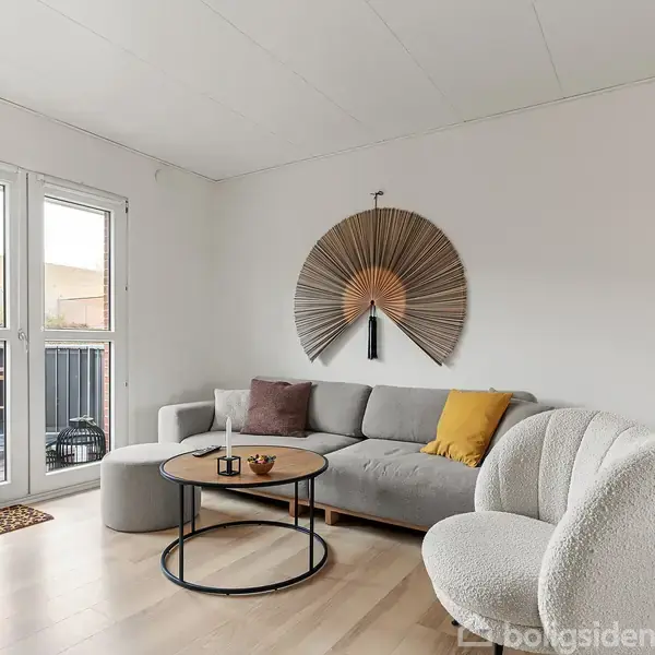 Gråt sofaarrangement med puder, sofabord i sort metal og træ. I baggrunden ses en stor vægdekoration. Rummet har en lys træbelægning og en altandør, der fører til en balkon.