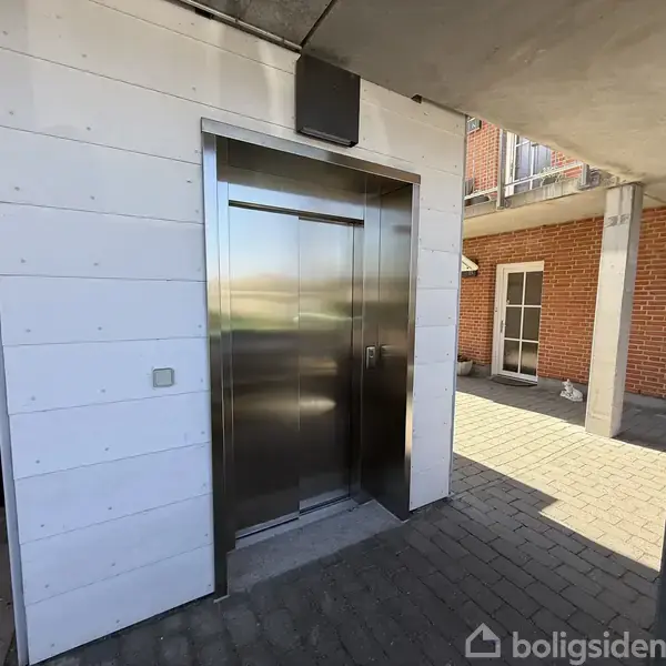 Elevator med metalfront står åben imellem hvide vægge. Den befinder sig i et udendørs gårdområde med brostensbelægning og røde murstensbygninger omkring.