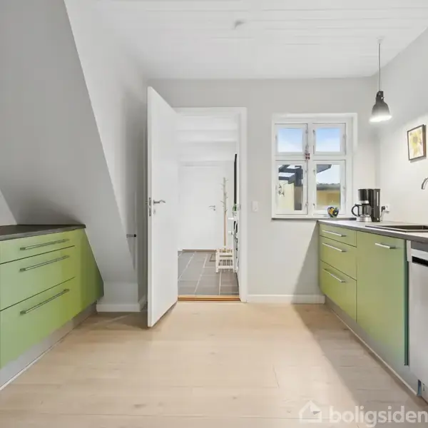 Køkken med grønne skabe på begge sider, indeholder køkkenredskaber. En åben dør fører til en gang; over vasken hænger lamper, og et vindue giver naturligt lys.