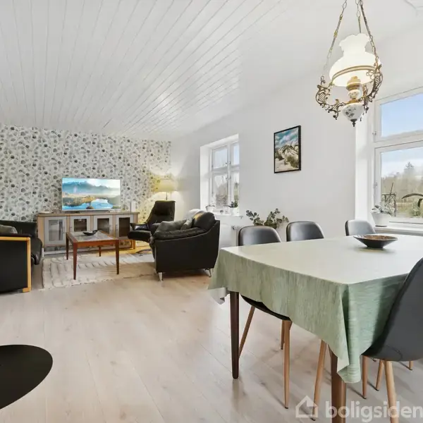 Stue med grøn dugklædt spisebord, sorte stole og brændeovn. Sofa og lænestole i hjørne foran tv og mønstret tapet. Store vinduer giver lys og udsigt til grønne omgivelser.