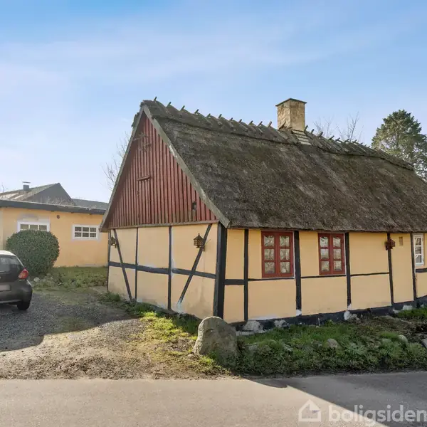 Bindingsværkshus med stråtag står stille på en åben, landlig grund. Ved siden af huset holder en sort bil parkeret på en grusvej. Himlen er klar og blå.