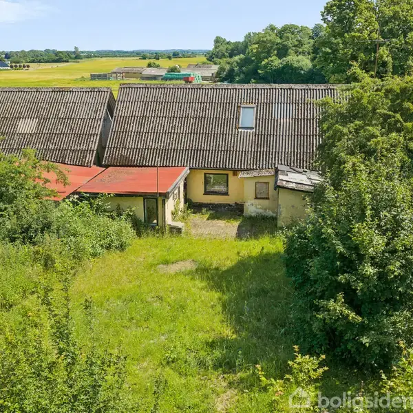 En ældre gårdbygning med et skråt tag, omgivet af tæt bevoksning. Bygningen står i en grøn landlig omgivelser med marker og træer i baggrunden.