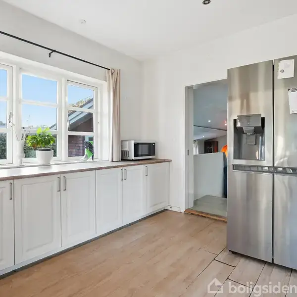 Køleskab står stille i et lyst køkken med hvide skabe. En kaffemaskine og mikroovn står på bordpladen. Kalender for 2026 hænger på køleskabet. Vindue med udsigt udenfor.