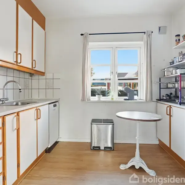 Et lyst køkken med hvide skabe og trækanter. Bordet står foran et vindue med udsigt til en have. Hylder holder diverse køkkenredskaber og en skraldespand står ved væggen.