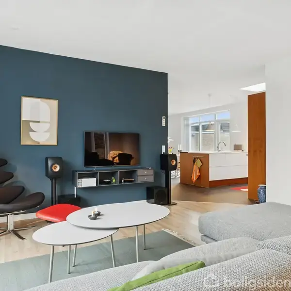 Stue med grå sofa, lænestol og sofabord foran en mørkeblå væg, hvor et fladskærms-TV hænger. I baggrunden ses åbent køkken med hvidt interiør og trædetaljer.