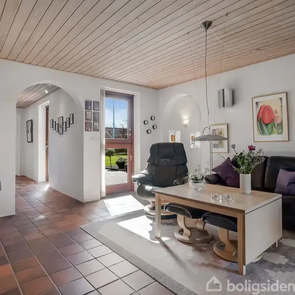 En stue med to lænestole og et sofabord, placeret på et tæppe. En brændeovn står til venstre. Sollys stråler ind gennem en dør mod en frodig have.