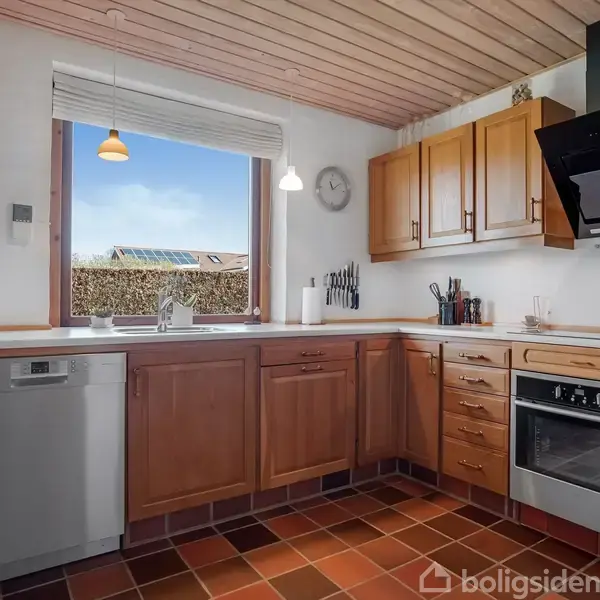 Et køkken med træskabe, der har opvaskemaskine, vask og komfur. Køkkenet har et stort vindue med udsigt til en hæk og flisegulv med rødlige nuancer.