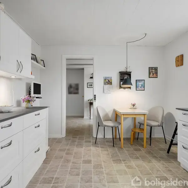 Et lyst køkken med hvide skabe og sorte bordplader. Et lille spisebord med to stole står ved en væg med billeder. Vindue til højre giver naturligt lys.