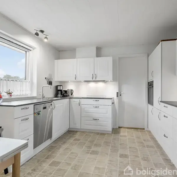 Et lyst køkken med hvide skabe og skuffer. En kaffemaskine står på bordpladen. En vindueskarm med dekorationer er synlig. Bordet er delvist synligt i forgrunden.