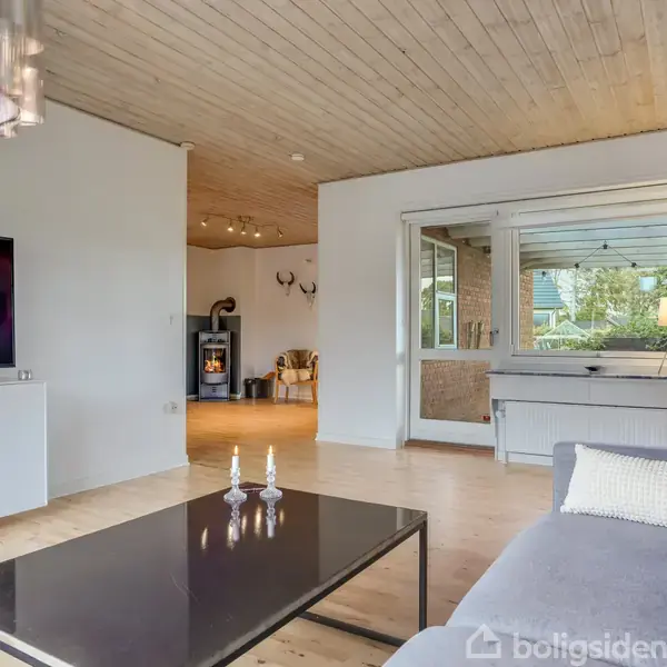 Stue med grå sofa og sort sofabord i forgrunden. Et fjernsyn står på en hvid skænk til venstre. I baggrunden ses et vindue og et rum med en pejs.