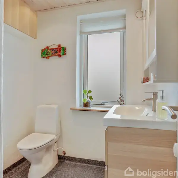 Toilet med hvidt sæde står i et lyst badeværelse. En lille plante og et dekorativt element pynter vindueskarmen. Skabe og håndvask er til højre.
