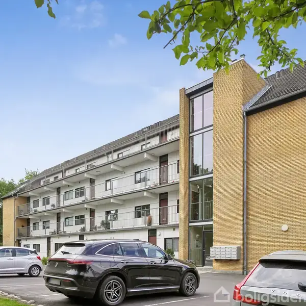 Et gult murstensbyggeri med altaner strækker sig over tre etager. Biler er parkeret foran bygningen, omgivet af grønne træer, under en klar blå himmel.