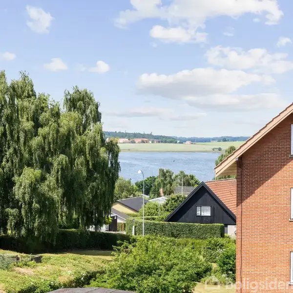 Rødt murstenshus står stille i forgrunden, omgivet af frodige grønne træer; i baggrunden ses en rolig sø under en klar, blå himmel.