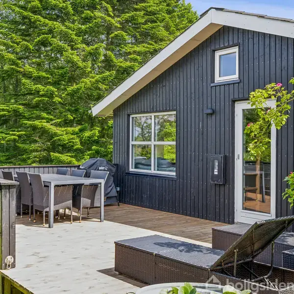 Sort træhus med terrasse, udstyret med spisebord og stole. Omgivet af grønne træer, under en klar blå himmel, skaber en fredelig atmosfære.
