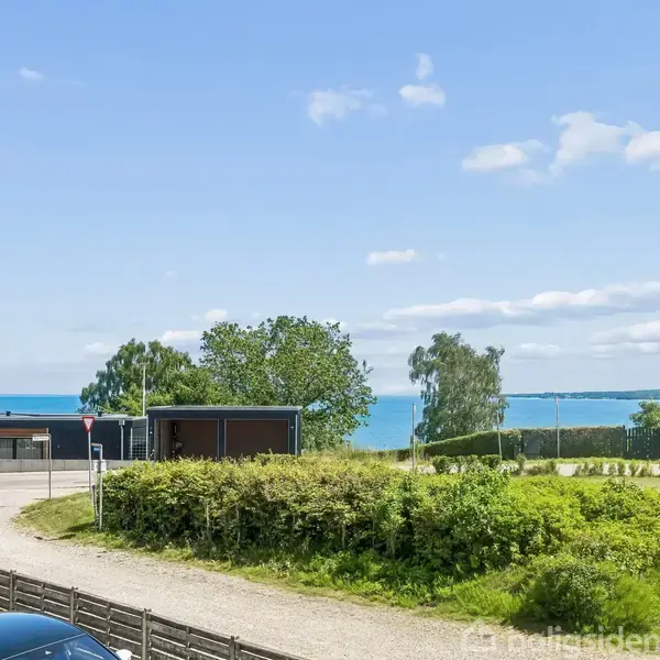 Moderne hus med fladt tag står stille i en idyllisk, grøn have tæt på en grusvej. Havudsigt og blå himmel danner en rolig baggrund.