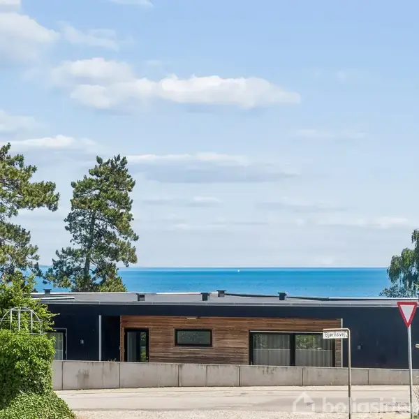 Et moderne, lavt hus står stille foran havet, omkranset af grønne træer og en klar, blå himmel. Området omkring er grønt og byder på en rolig udsigt. Skiltet viser "Bekkevang."