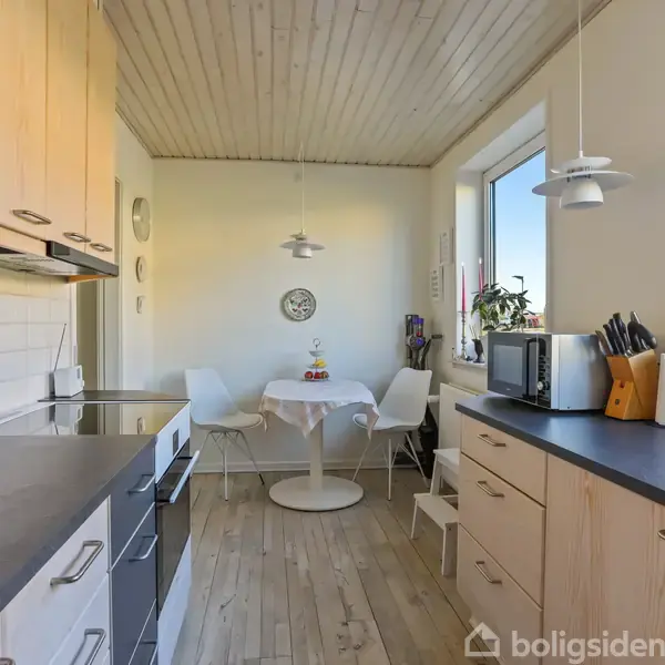 Køkken med træskabe og sort bordplade. Rundt bord med stole i hjørne, pyntet med planter og køkkenredskaber. Trægulv og lyse vægge skaber en lys og hyggelig atmosfære.