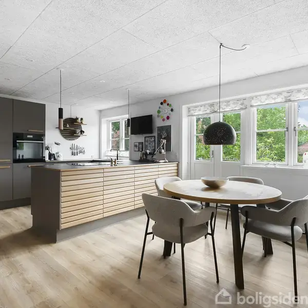 Moderne køkken med gråt skab og træbordplade, midt i madlavning, omgivet af store vinduer med udsigt til grønne træer og en spisestue med rundt bord og grå stole.