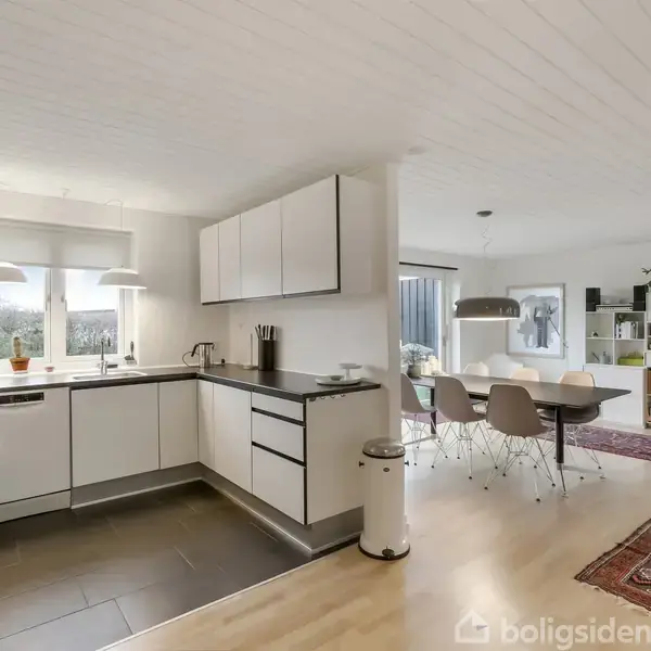 Et moderne hvidt køkken er placeret i et lyst rum med to vinduer. I nærheden står et spisebord med seks stole og en reol i hjørnet på et tæppe.