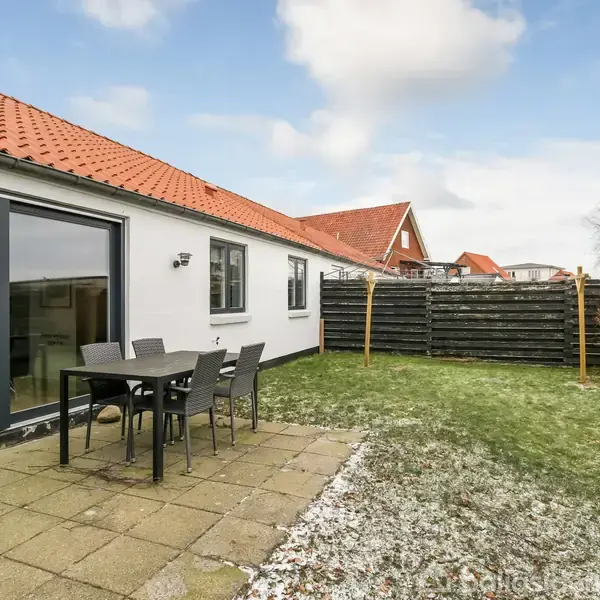 Et bord med fire stole står på en flisebelagt terrasse ved siden af et hvidt hus med store vinduer. I baggrunden ses en græsplæne og et plankeværk.