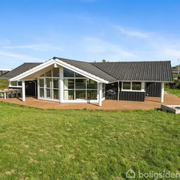 Et moderne sommerhus står på en stor, grøn græsplæne. Husets facade har store vinduer, der vender mod en åben terrasse. Huset er omgivet af et landskab med flere sommerhuse i baggrunden.