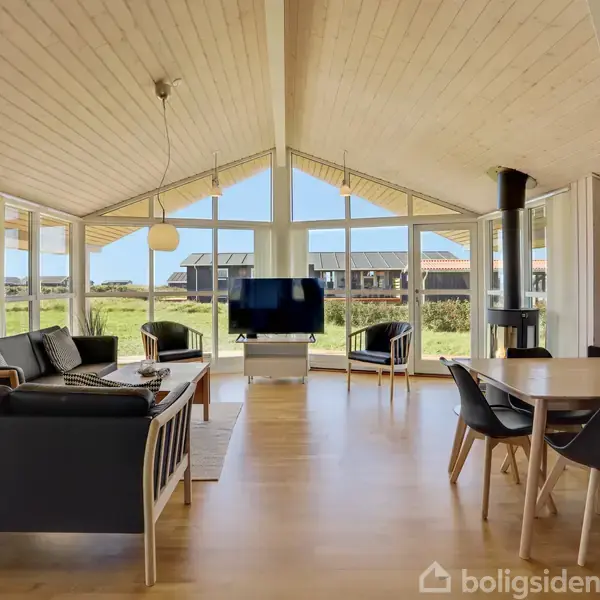 Stue med sorte sofaer og fladskærms-TV, beliggende i et rum med højt til loftet og store vinduer. Langbord med moderne stole, omkranset af lys og udsigt til grønne omgivelser.
