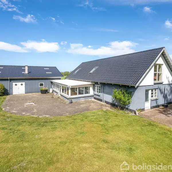 Hus med sort tag ligger i en grøn have. Tilknyttet garagebygning på venstre side. Blå himmel med skyer i baggrunden.