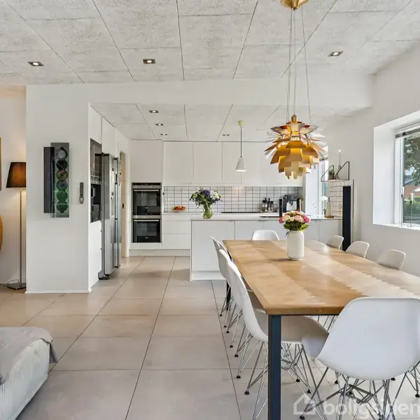 Spisebord med hvide stole står i et lyst køkkenalrum. Rummet har store vinduer, hvidt interiør og en gul lænestol. En moderne lampe hænger over bordet, mens planter pynter vindueskarmen.