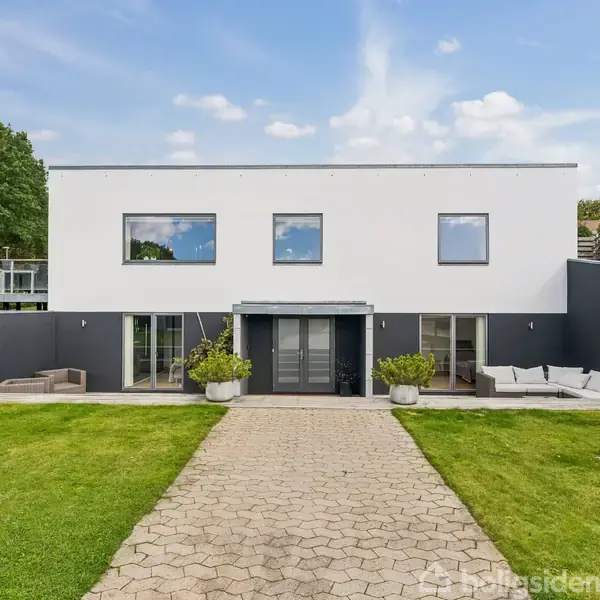 En moderne to-etagers bygning står på en velplejet græsplæne. Bygningen har store glasvinduer og en stenbelagt sti, der fører op til hovedindgangen. To terrasseområder ses på hver side.