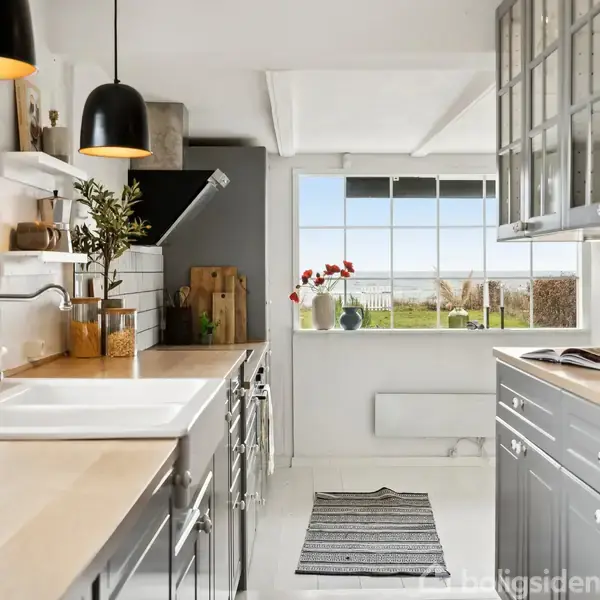 Køkken med grå skabe og lyst træbordplade. Store vinduer viser en have. Hængelamper og planter pynter rummet. Skærebrætter og glasbeholdere er synlige på bordet.