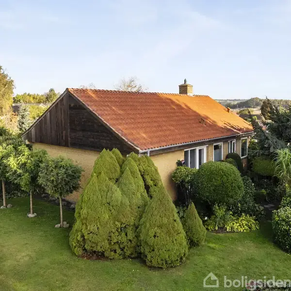 Et hus med rød tag er omgivet af en velplejet have med frodige træer og buske. Det ligger i et landskabeligt område med udsigt til en skov i baggrunden.