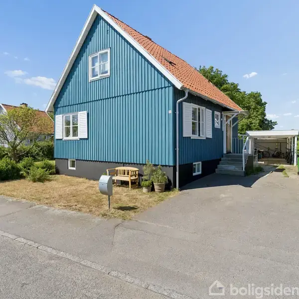 Blåt træhus med rødt tag står på en stille villavej. Foran huset er en lille have med græs og buske. Tilstødende er en indkørsel og carport.