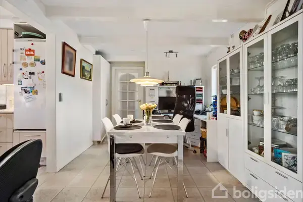 Et spisebord med stole står i centrum, dekoreret med en vase med blomster. Omkring bordet er der hvide skabe fyldt med glas og service. Kjøkken og kontor er synligt i baggrunden.