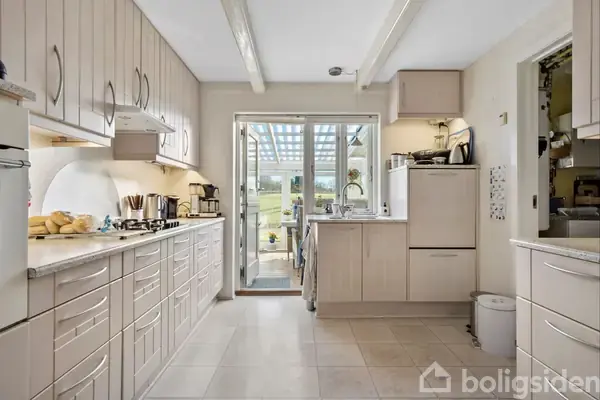 Et lyst køkken med hvide skabe og skuffer rummer en kogeplade med gryder og køkkenudstyr. En glasdør fører ud til en overdækket terrasse med udsigt til en have.