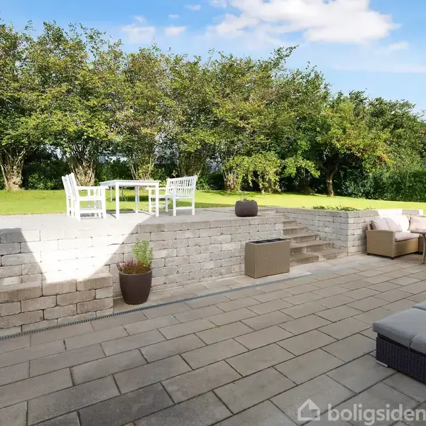 Terrasse med loungeområde, hvor møbler inkluderer sofa og bord, står på flisebelægning. Øverst er en hævet græsplæne med et sæt havemøbler. Baggrunden har tætte grønne træer.