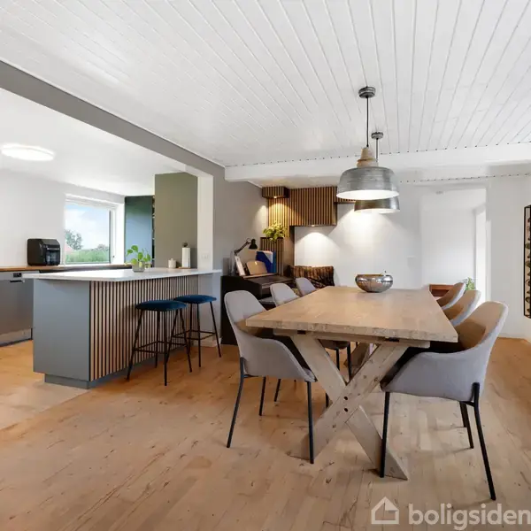 Spisebord i træ med seks stole er omdrejningspunktet i et moderne køkken-alrum. Store lamper hænger over bordet, og i baggrunden ses en køkkenø med barstole.