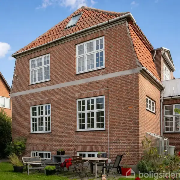 Et rødt murstenshus med hvide sprossevinduer, omgivet af en grøn have med havemøbler. Huset har en rød tegltag, og der er en klar blå himmel i baggrunden.