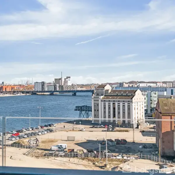Vandet strækker sig roligt foran en by, hvor hvide bygninger står tæt ved kajen. Biler er parkeret langs en grusvej. Skyer driver hen over himlen.